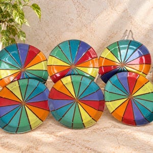 May include: A set of five round ceramic plates featuring a vibrant, multi-colored radial pattern. Each plate displays alternating triangular segments in shades of red, orange, yellow, green, blue, and purple, separated by thin dark lines.