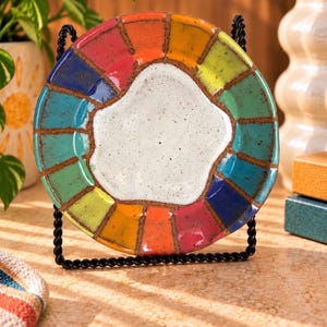 May include: A round ceramic plate featuring a central white speckled area surrounded by a border of multicolored rectangular segments in shades of red, orange, yellow, green, and blue, all separated by dark brown grout lines on a black stand.