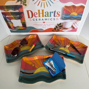 May include: Three rectangular ceramic trays with wavy edges, each featuring a hand-painted sunset design in orange, yellow, and teal. The trays hold various personal items including sunglasses, a lip balm, keys, tweezers, a pen, a ring, and gum.