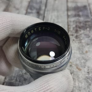 Pu&ograve; includere: Un obiettivo per fotocamera vintage con il testo "ЮПИТЕР-3" e altri segni. L'obiettivo ha un corpo in metallo nero e argento con un elemento in vetro. L'obiettivo &egrave; tenuto da una mano guantata.