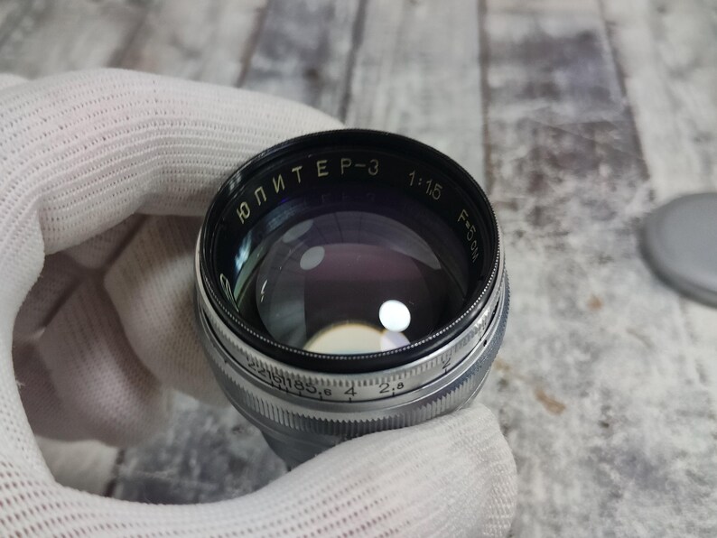 Pu&ograve; includere: Un obiettivo per fotocamera vintage con il testo "ЮПИТЕР-3" e altri segni. L'obiettivo ha un corpo in metallo nero e argento con un elemento in vetro. L'obiettivo &egrave; tenuto da una mano guantata.