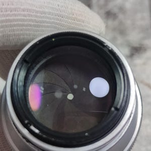 Pu&ograve; includere: Primo piano di un obiettivo fotografico con corpo in metallo argentato. L'interno dell'obiettivo &egrave; scuro, con lamelle che formano un'apertura. Un riflesso bianco brillante &egrave; visibile, insieme a una sfumatura rosa e viola.