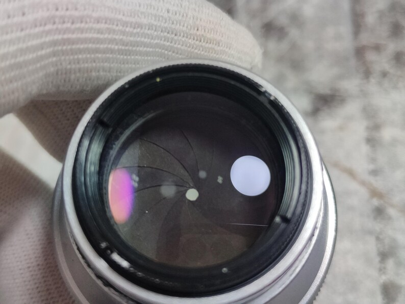 Pu&ograve; includere: Primo piano di un obiettivo fotografico con corpo in metallo argentato. L'interno dell'obiettivo &egrave; scuro, con lamelle che formano un'apertura. Un riflesso bianco brillante &egrave; visibile, insieme a una sfumatura rosa e viola.