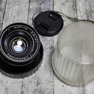 May include: A black camera lens with the text "МИР-1Ш 2,8/37" and serial number "8802088N". Includes a black lens cap and a clear plastic lens case. The lens is on a wooden surface.