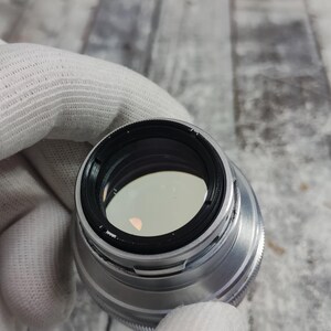 Pu&ograve; includere: Un obiettivo per fotocamera argentato con interno nero ed elemento in vetro trasparente. L'obiettivo &egrave; tenuto da una mano guantata, con un tappo grigio visibile sullo sfondo. L'obiettivo ha una ghiera di messa a fuoco zigrinata.