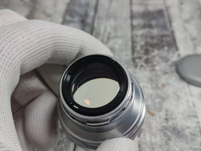 Pu&ograve; includere: Un obiettivo per fotocamera argentato con interno nero ed elemento in vetro trasparente. L'obiettivo &egrave; tenuto da una mano guantata, con un tappo grigio visibile sullo sfondo. L'obiettivo ha una ghiera di messa a fuoco zigrinata.