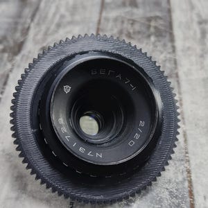 Puede incluir: Un objetivo de cámara negro con el texto "VEGA 7-1" y "2/20" impreso. El objetivo tiene un anillo exterior estriado y un borde dentado. El objetivo está sobre una superficie de madera gris.