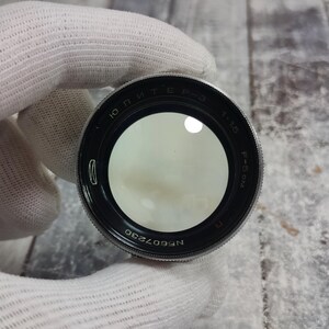 Pu&ograve; includere: Un obiettivo per fotocamera nero e argento con il testo "ЮПИТЕР P-3 1:1.5 F=5cm" e il numero di serie "N607230". L'obiettivo &egrave; tenuto da una mano guantata. Un tappo per obiettivo argentato &egrave; visibile sullo sfondo.