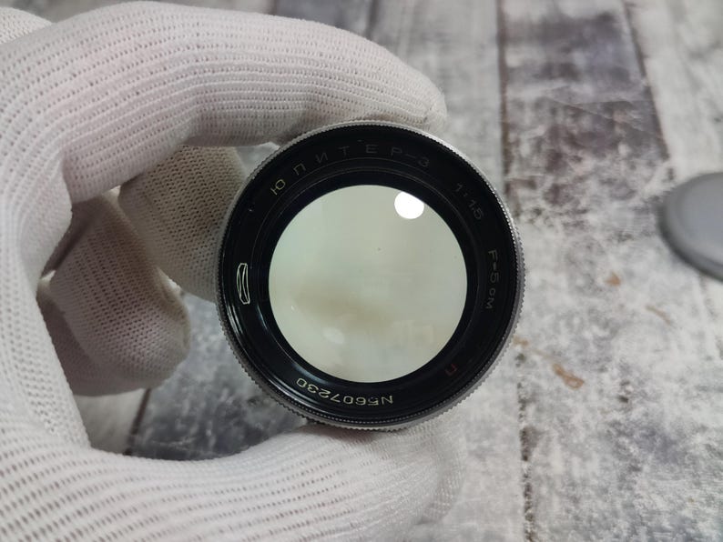 Pu&ograve; includere: Un obiettivo per fotocamera nero e argento con il testo "ЮПИТЕР P-3 1:1.5 F=5cm" e il numero di serie "N607230". L'obiettivo &egrave; tenuto da una mano guantata. Un tappo per obiettivo argentato &egrave; visibile sullo sfondo.
