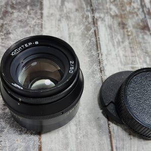 May include: A black camera lens with the text "ЮПИТЕР-8" and "2/50" printed on the lens. The lens is accompanied by two black lens caps. The lens is on a wooden surface.