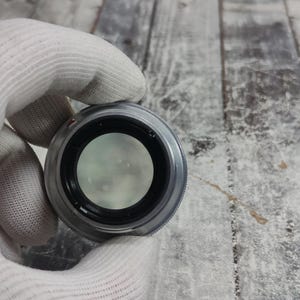 Pu&ograve; includere: Un obiettivo per fotocamera argentato con interno nero e vetro trasparente. L'obiettivo &egrave; tenuto da una mano guantata. Un tappo dell'obiettivo grigio &egrave; visibile sullo sfondo. L'obiettivo &egrave; progettato per la fotografia.