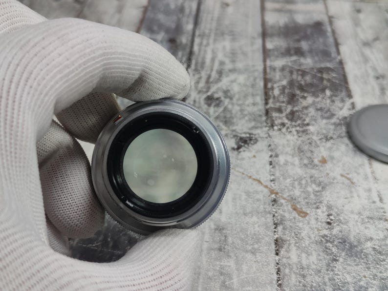 Pu&ograve; includere: Un obiettivo per fotocamera argentato con interno nero e vetro trasparente. L'obiettivo &egrave; tenuto da una mano guantata. Un tappo dell'obiettivo grigio &egrave; visibile sullo sfondo. L'obiettivo &egrave; progettato per la fotografia.