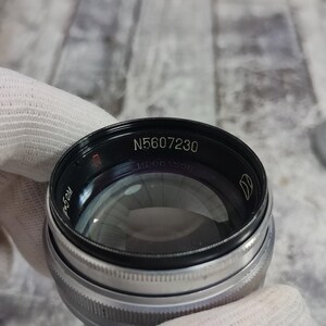 Pu&ograve; includere: Primo piano di un obiettivo fotografico con la scritta "N5607230" in bianco. L'obiettivo &egrave; nero e argento, con un elemento in vetro trasparente. L'obiettivo &egrave; tenuto da una mano guantata.
