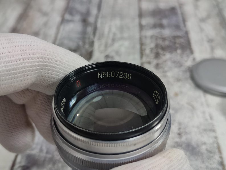 Pu&ograve; includere: Primo piano di un obiettivo fotografico con la scritta "N5607230" in bianco. L'obiettivo &egrave; nero e argento, con un elemento in vetro trasparente. L'obiettivo &egrave; tenuto da una mano guantata.