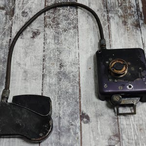 Op de afbeelding: Een antiek, zwart en paars apparaat met een gebogen, donkere kabel die twee componenten verbindt. Eén component is een rechthoekige doos met een ronde wijzerplaat en schroeven. De andere is een afgerond, plat stuk. Het apparaat ligt op een verweerd, houten oppervlak.