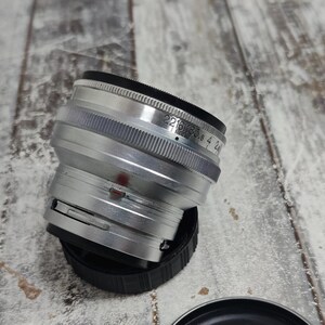 Pu&ograve; includere: Un obiettivo per fotocamera argentato con dettagli neri e un tappo nero. L'obiettivo presenta un anello di messa a fuoco zigrinato e contrassegni di apertura. L'obiettivo &egrave; posizionato su una superficie di legno.