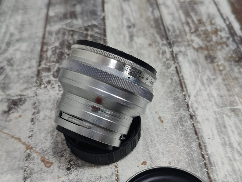 Pu&ograve; includere: Un obiettivo per fotocamera argentato con dettagli neri e un tappo nero. L'obiettivo presenta un anello di messa a fuoco zigrinato e contrassegni di apertura. L'obiettivo &egrave; posizionato su una superficie di legno.