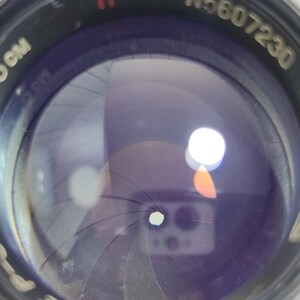 Pu&ograve; includere: Primo piano di un obiettivo fotografico con un involucro in metallo nero. L'obiettivo ha lamelle interne e una superficie riflettente. Il testo "F=5 cm" e "1:1.5" &egrave; visibile.