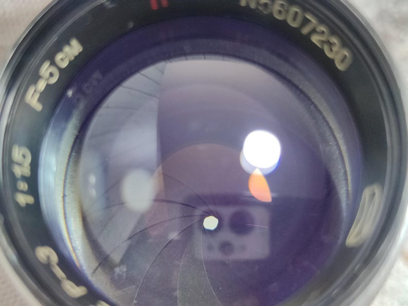 Pu&ograve; includere: Primo piano di un obiettivo fotografico con un involucro in metallo nero. L'obiettivo ha lamelle interne e una superficie riflettente. Il testo "F=5 cm" e "1:1.5" &egrave; visibile.