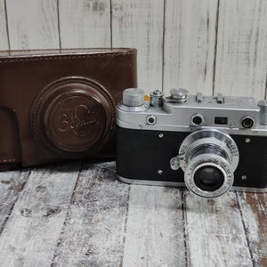 Puede incluir: Una cámara plateada y negra vintage con lente y diales, junto a una funda de cuero marrón. La funda tiene un emblema circular. La cámara está sobre una superficie de madera.