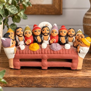May include: A terracotta-colored clay sculpture depicting a group of thirteen figures seated at a long rectangular table. The figures wear colorful pointed hats and white garments, with small clay food items placed on the table surface in front of them.