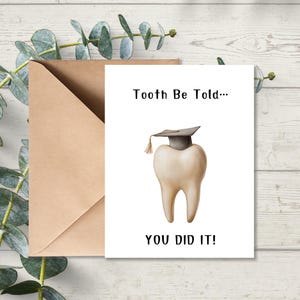 May include: A greeting card with a tooth wearing a graduation cap. The card reads "Tooth Be Told... YOU DID IT!" and is accompanied by a brown envelope. The background is a light wood surface with green foliage.