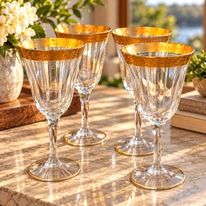 May include: Four clear glass goblets featuring vertical fluted patterns and wide gold rims with intricate etched detailing. Each glass has a slender stem and a circular base with a thin gold edge, reflecting warm light on the surface.