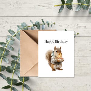 May include: A birthday card with a watercolor illustration of a squirrel holding a cake with lit candles. The card reads "Happy Birthday" in black text. A brown envelope is behind the card, with eucalyptus leaves in the background.