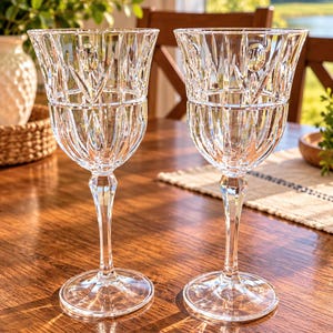 May include: Two clear glass goblets featuring a detailed cut-glass pattern on the bowl and a faceted stem. The design includes vertical lines and circular accents around the upper rim, supported by a circular base for stability and balance.