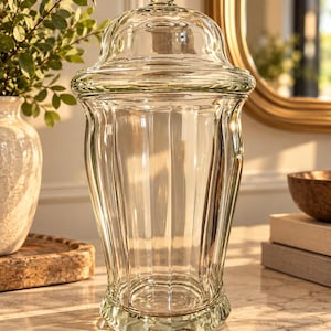 May include: A clear glass apothecary jar with a matching lid featuring a spherical finial. The jar has a vertical ribbed design, a wide flared rim, and a tapered base, creating a tall, elegant silhouette for storage or display purposes.