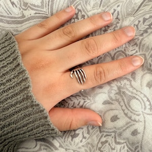 May include: A silver skeleton hand ring. The ring is worn on the middle finger of a hand.