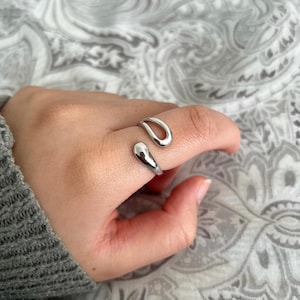 May include: A silver ring with a unique, abstract design. The ring is open and wraps around the finger.