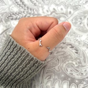 May include: Two silver rings, one on the thumb and the other on the index finger. The rings feature a minimalist design with a small, abstract shape. The rings are worn on a hand, with a grey sweater visible in the background.