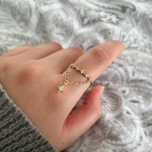 May include: A gold chain ring with a dangling star charm. The ring is adorned with small clear cubic zirconia stones.