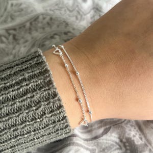 May include: Two silver bracelets on a wrist. One bracelet has a delicate chain with small silver beads. The other bracelet has a thicker chain with a heart charm.