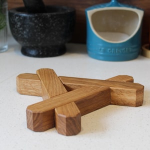 May include: A wooden trivet with a geometric design. The trivet is made of light brown wood and has a cross-like structure. It is designed to protect surfaces from hot pots and pans. Kitchen decor.