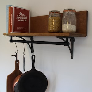 May include: A wooden kitchen shelf with a black metal rod and hanging hooks. The shelf holds two glass jars with food, cookbooks, and kitchen utensils. A wooden chopping board, a copper pan, and a black cast iron frying pan hang below.