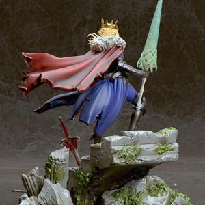 May include: A detailed figure of a fantasy character wearing a blue and red outfit with a gold crown and holding a spear. The figure is standing on a rocky base with green moss.