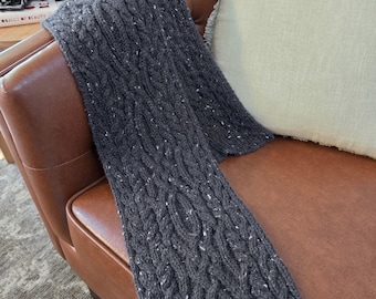 May include: A dark charcoal gray knit scarf featuring a prominent cable knit pattern with small white flecks throughout the yarn. The scarf is draped over a brown leather surface next to a light green pillow and vintage cameras.