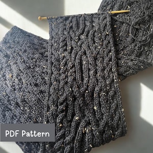 May include: Dark gray knitted scarf with a cable knit design. The scarf is made of a speckled yarn and is partially on knitting needles. The text "PDF Pattern" is in a gray box in the lower left corner.