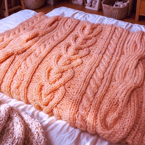 Könnte beinhalten: Eine pfirsichfarbene, grob gestrickte Decke mit Zopfmuster. Die Decke ist auf einem weißen Bett gefaltet und zeigt die komplizierte Textur und warme Farbe. Die Decke ist aus weichem, dickem Garn gefertigt.