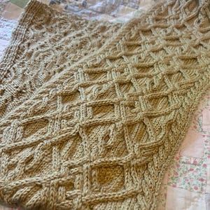 May include: A tan-colored, hand-knitted blanket with a geometric pattern. The blanket features a textured design with a combination of cable and lattice stitches. The soft, warm blanket is perfect for snuggling up on the couch or bed.