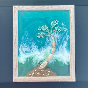 Könnte beinhalten: Gerahmtes Kunstwerk aus Kunstharz, das eine Palme an einem Strand zeigt. Der Baum besteht aus kleinen, hellen Muscheln, die vor einem türkisfarbenen und weißen Ozeanhintergrund angeordnet sind. Der Rahmen besteht aus hellem Holz.