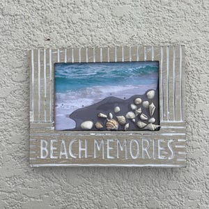 Può includere: Una cornice rettangolare in legno con la scritta "BEACH MEMORIES" in lettere bianche. La cornice contiene una foto di una scena di spiaggia con conchiglie. La cornice è di colore marrone chiaro con strisce bianche verticali.