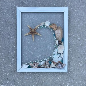 Può includere: Un'opera d'arte con cornice bianca con una stella marina e una disposizione di conchiglie, perle e piccole perline blu. Le conchiglie sono di varie forme e colori, creando un tema costiero.
