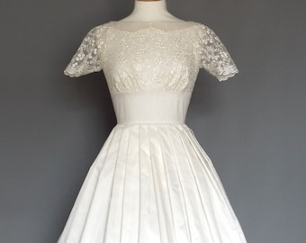 Puede incluir: Un vestido blanco vintage con un corpiño ajustado y una falda plisada amplia. El corpiño presenta delicadas mangas de encaje y un escote redondo. El vestido se exhibe en un maniquí de madera sobre un fondo gris. Un diseño clásico y elegante.