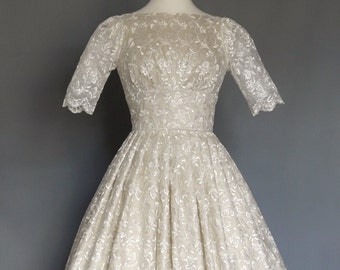 Puede incluir: Un vestido de novia de encaje blanco con un corpiño ajustado y una falda completa. El vestido tiene un escote redondo y mangas cortas. La falda está adornada con un delicado patrón floral.