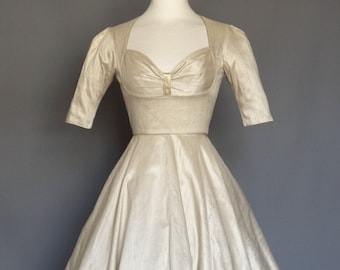 Puede incluir: Un vestido vintage de seda marfil con escote de corazón y falda completa. El vestido tiene un ribete dorado alrededor del escote y un lazo en el centro.