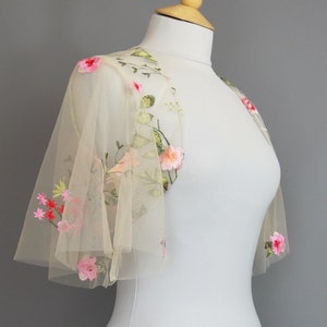 Peut inclure: Un petit capuchon beige transparent avec broderie florale. Le capuchon présente des fleurs brodées roses, rouges et vertes sur les épaules et les manches. Le vêtement est présenté sur un mannequin blanc sur fond gris.