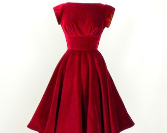 Puede incluir: Un vestido de terciopelo rojo con un corpiño ajustado y una falda completa. El vestido tiene un escote barco y mangas cortas.